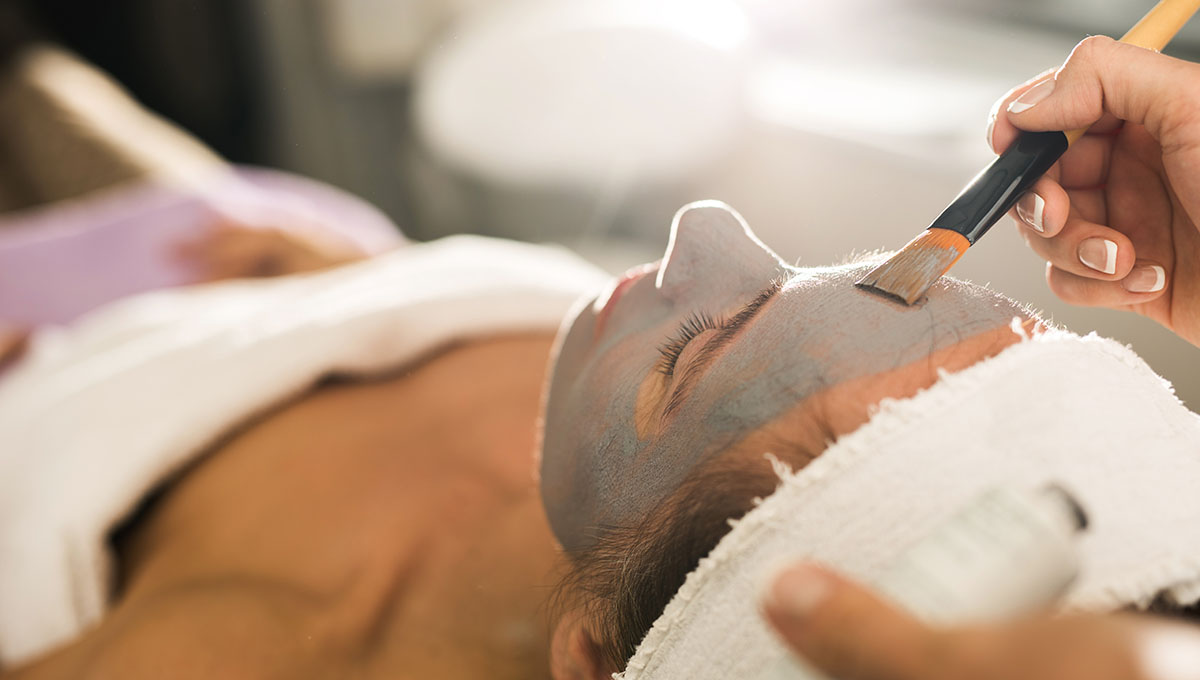 Close up of unrecognizable beautician applying clay mask on woman's face during beauty treatment.