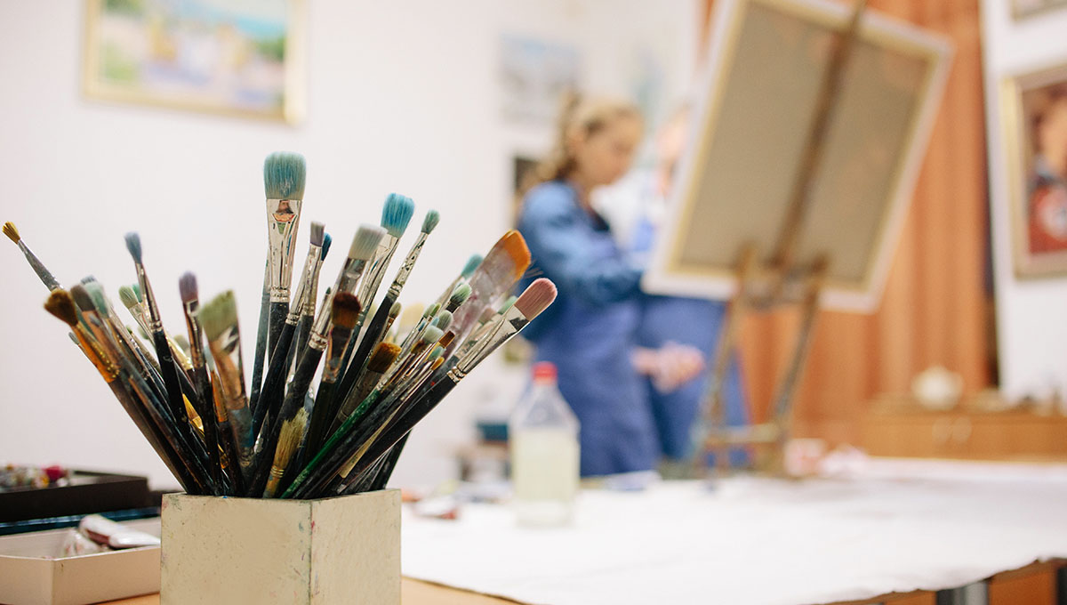 A close up shot of paint brushes in a pot while an artist paints on an easil in the background