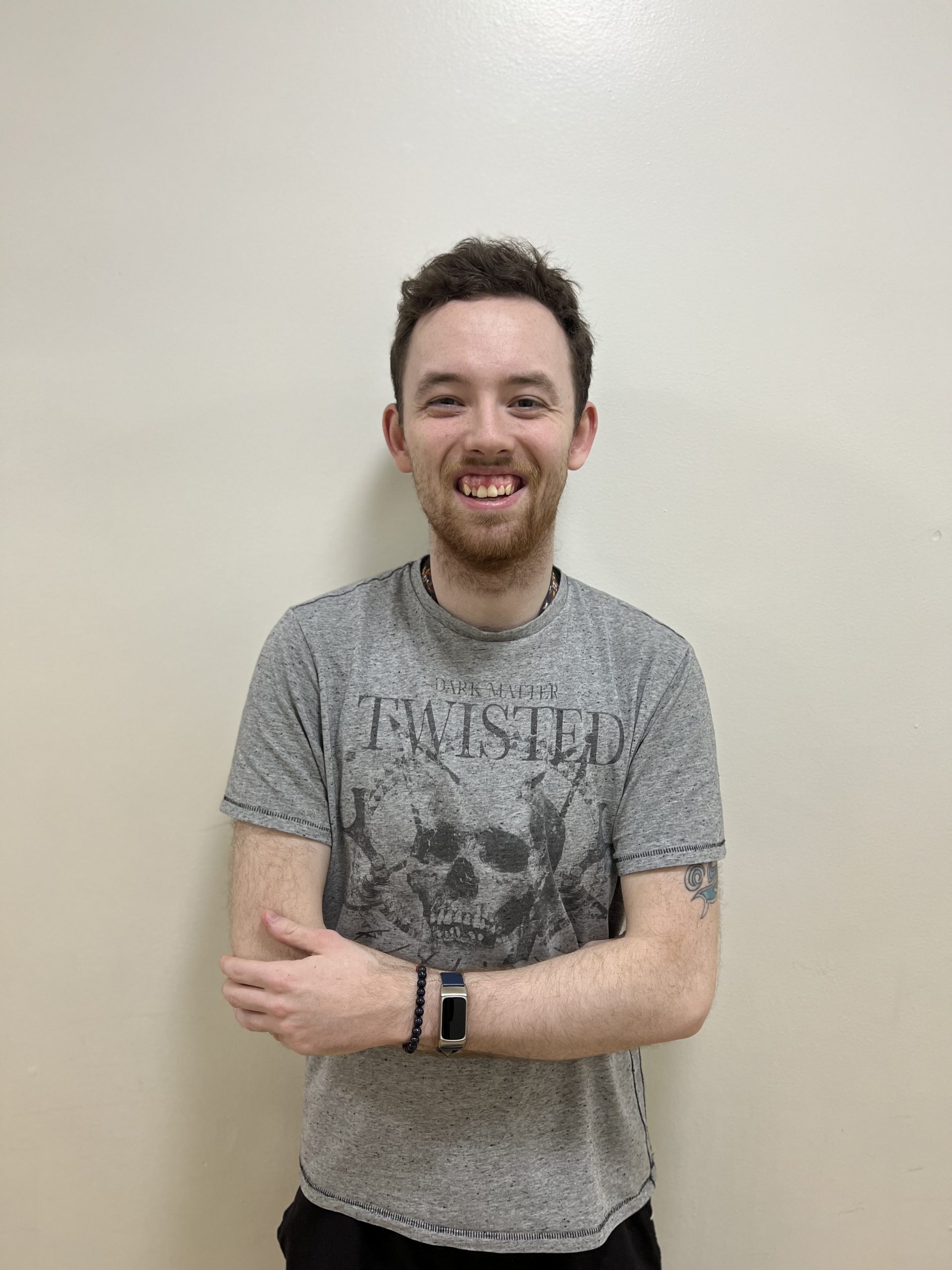 A young man stands against a wall smiling with his arms folded.