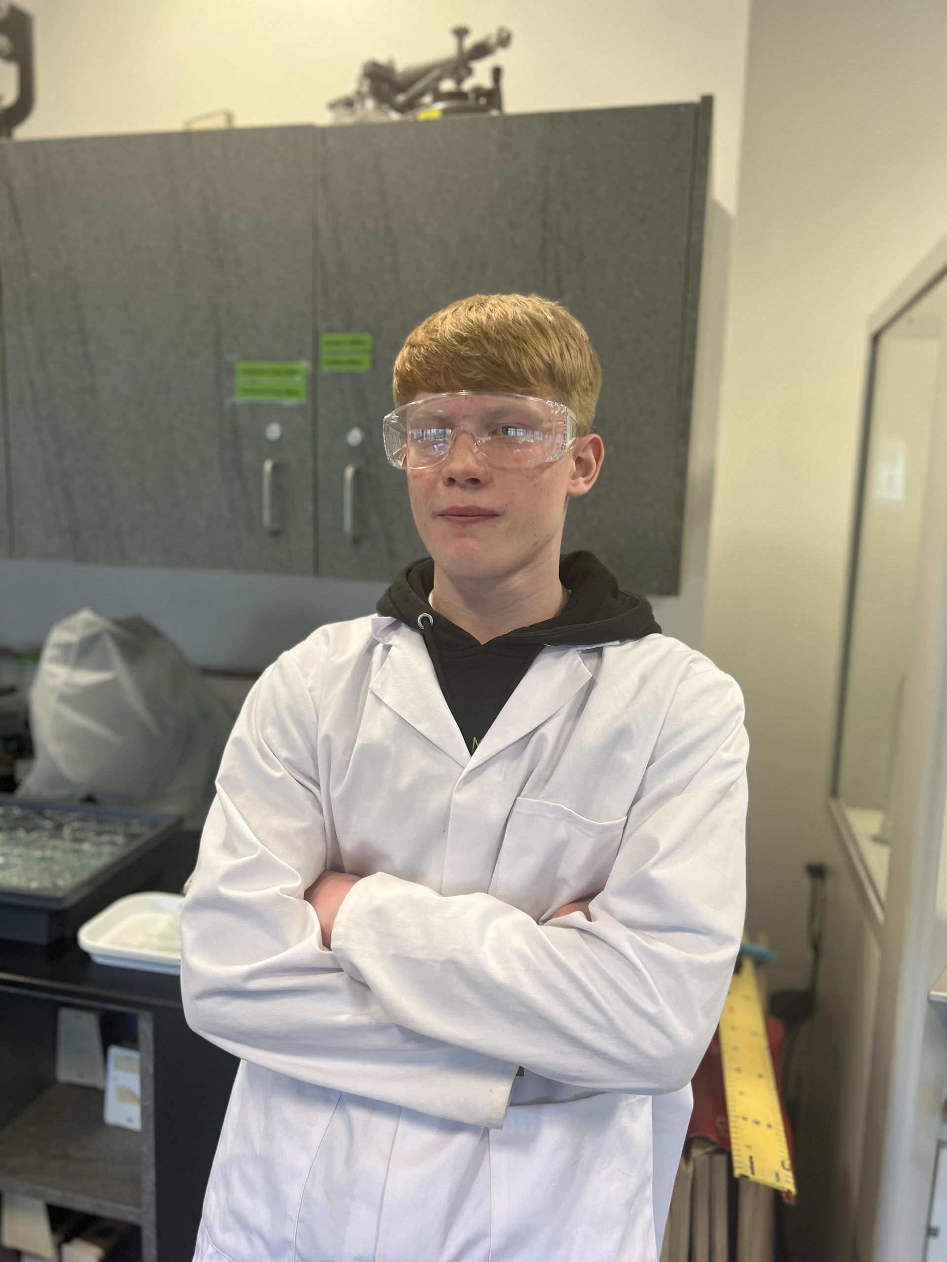 A young man stands in a lab wearing a lab coat and protective goggles.