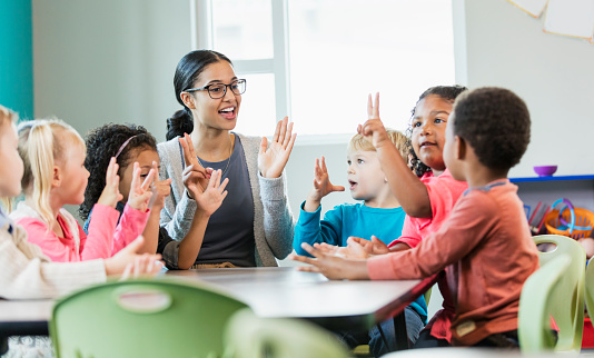 A multi-ethnic group of six preschool children with a mixed race African-American and Caucasian teacher, sitting around a table in a classroom. The teacher and some of her students have their hands raised, holding up fingers, learning how to count.