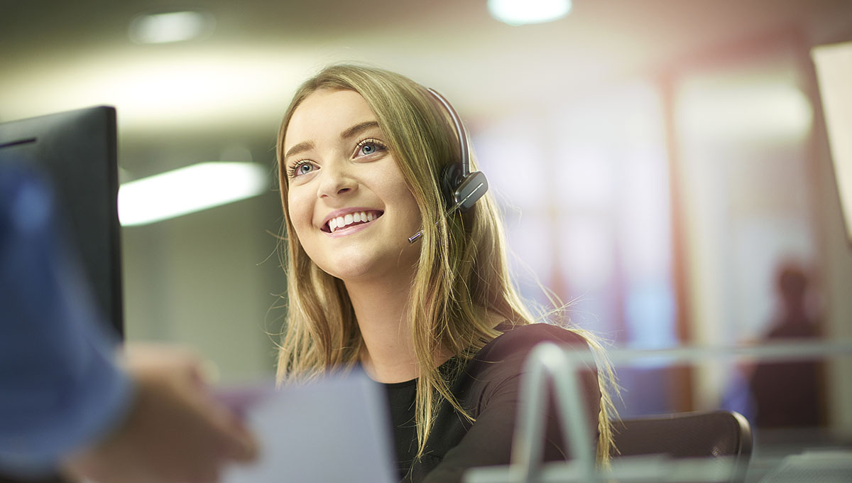 a young woman sits at her desk and is handed some paperwork by a colleague out of shot. She is smiling and enjoying her placement at the large open plan office . She is wearing a headset .