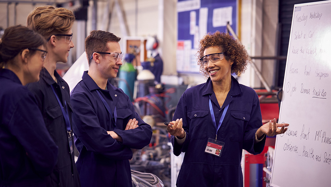 A woman teacher laughing with three male students in a workshop all wearing overalls