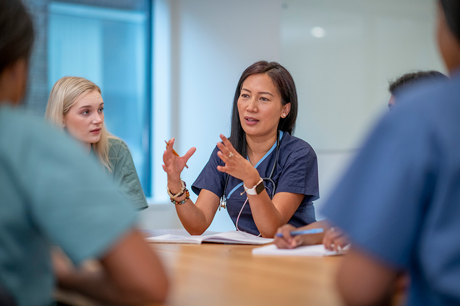 An asian lady in scrubs talks to a group of people all sitting at table.